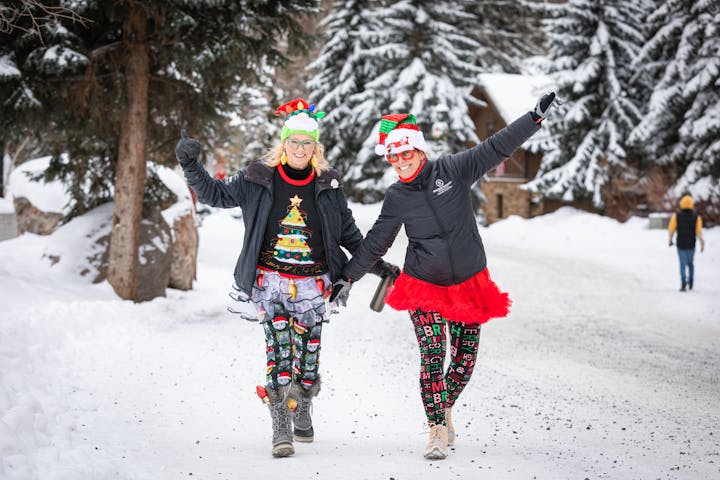 Two people in festive outfits and hats walking in a snowy landscape, smiling and having fun.