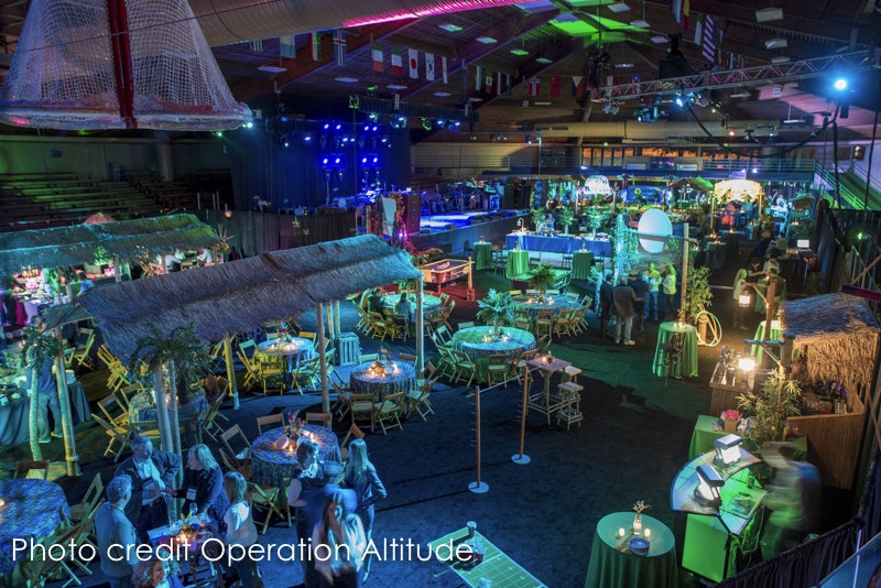 A tropical-themed event with thatched huts, tables, and a stage with blue lights, indoors. People are mingling.