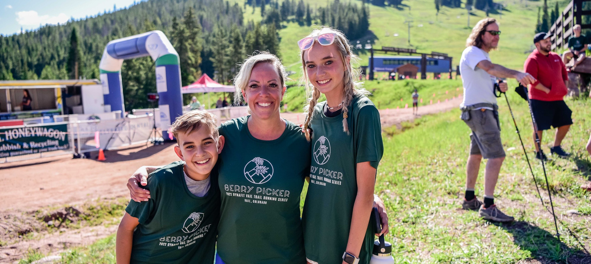 Three people smiling in matching shirts, at an outdoor event with running race finish line and spectators in the background.