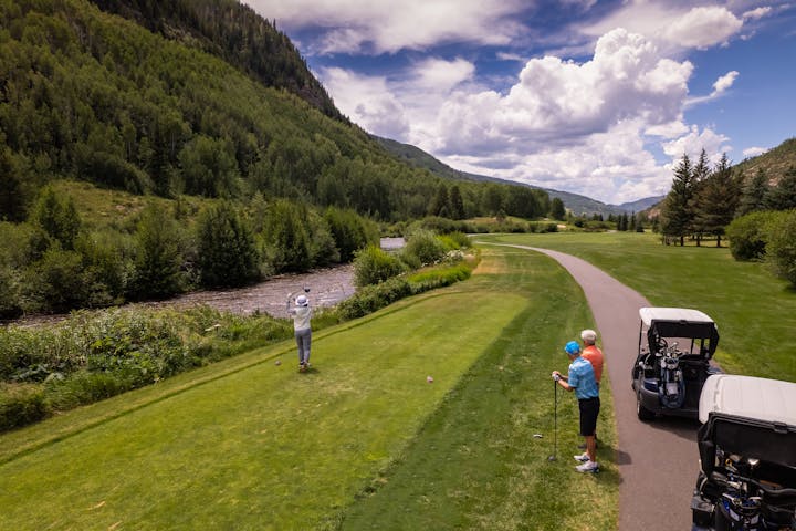 A golfer swinging a club, an observer, golf cart, and a scenic landscape with trees and a river.