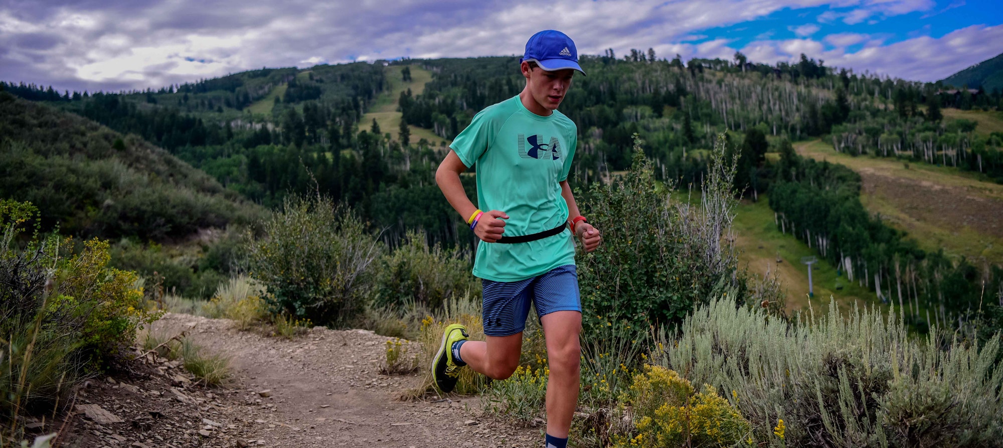 A person running on a mountain trail with scenic green hills and trees in the background.