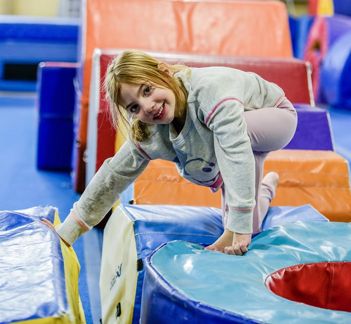 A child playing on colorful padded gym equipment indoors, smiling at the camera.
