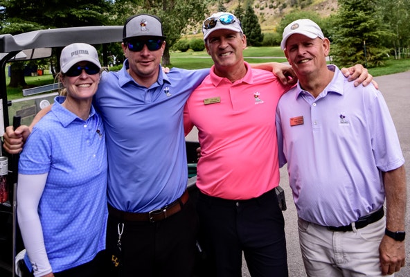 Four individuals smiling at a golf course, three men and one woman, dressed in golf attire.