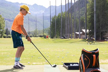 A kid at a driving range practicing golf, with a club in hand and a bag of golf balls nearby.