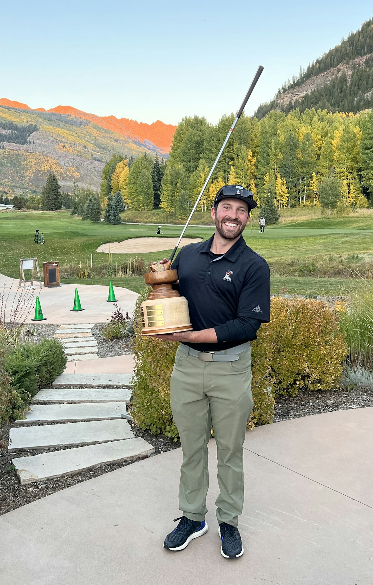 Man holding trophy and golf club, smiling, with mountains and golf course in the background.