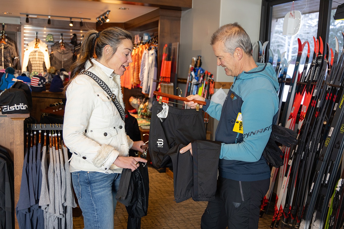 Two people in a ski shop, one holding ski pants, surrounded by clothes and ski gear.