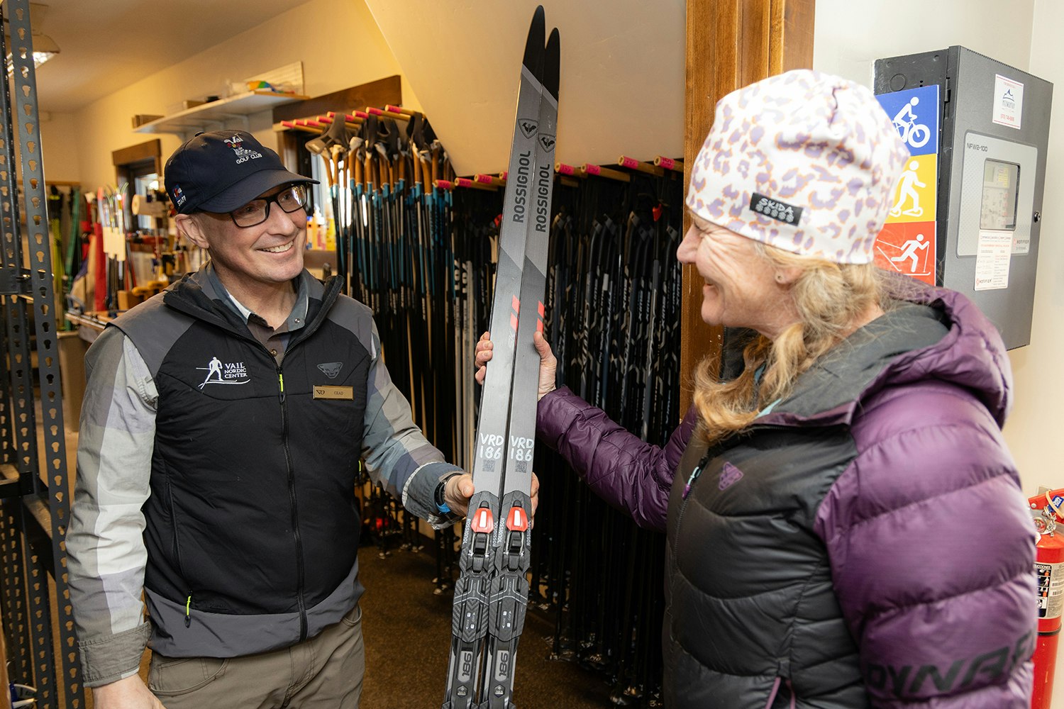 Two people smiling and holding cross-country skis in a ski rental shop.