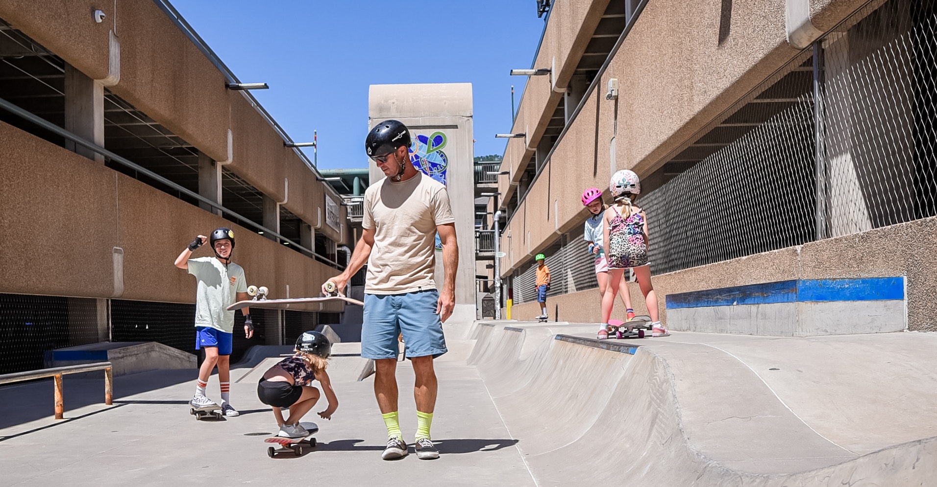 A group of people with skateboards at a skatepark, including a male instructor and children wearing helmets.