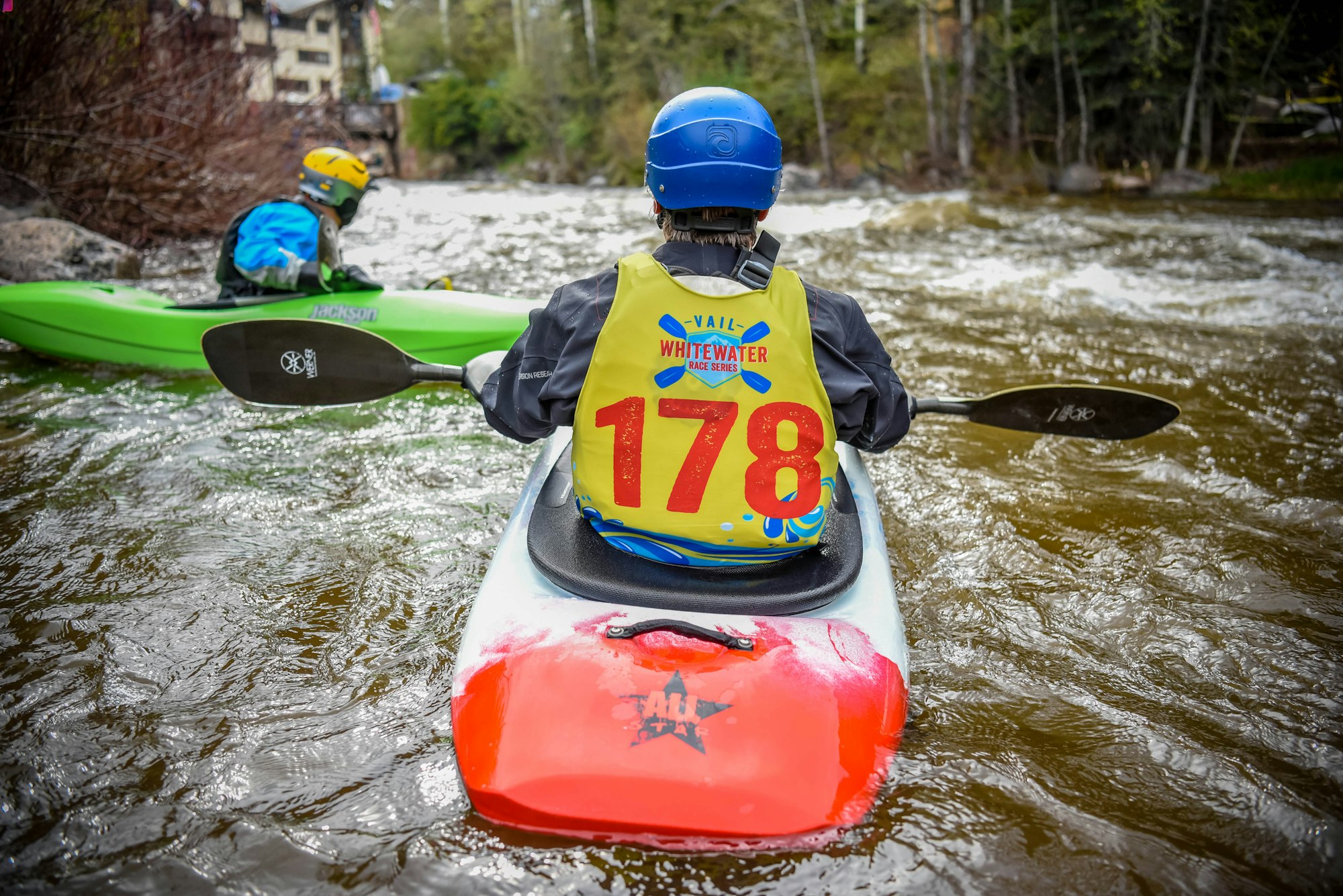 Two kayakers wearing helmets and life vests numbered "178" preparing for a whitewater race on a river.