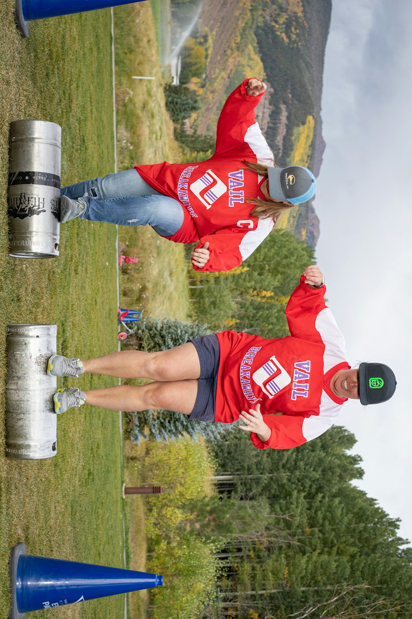 Two people in jerseys balancing on kegs outdoors, with a mountain landscape in the background.