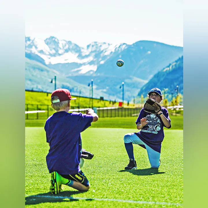 Two kids playing catch with a baseball on a sunny field with mountains in the background.