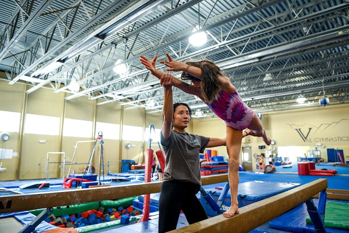A coach assisting a young gymnast on the balance beam in a gym.
