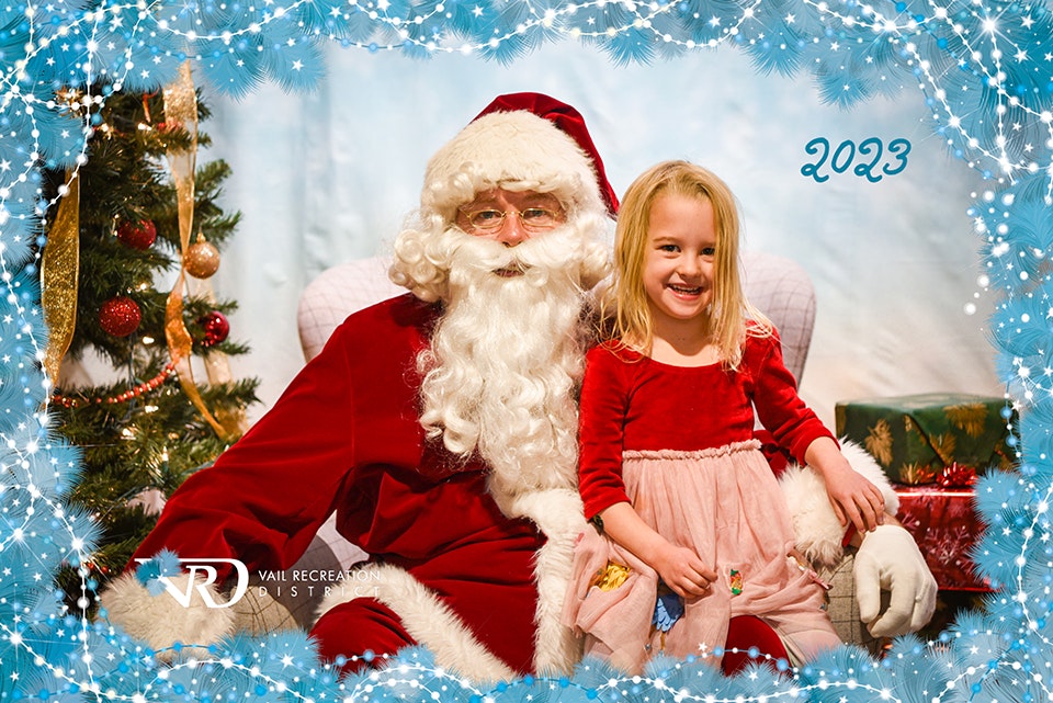 A person dressed as Santa Claus with a child, Christmas tree in background, photo framed with "2023" and snowflakes.