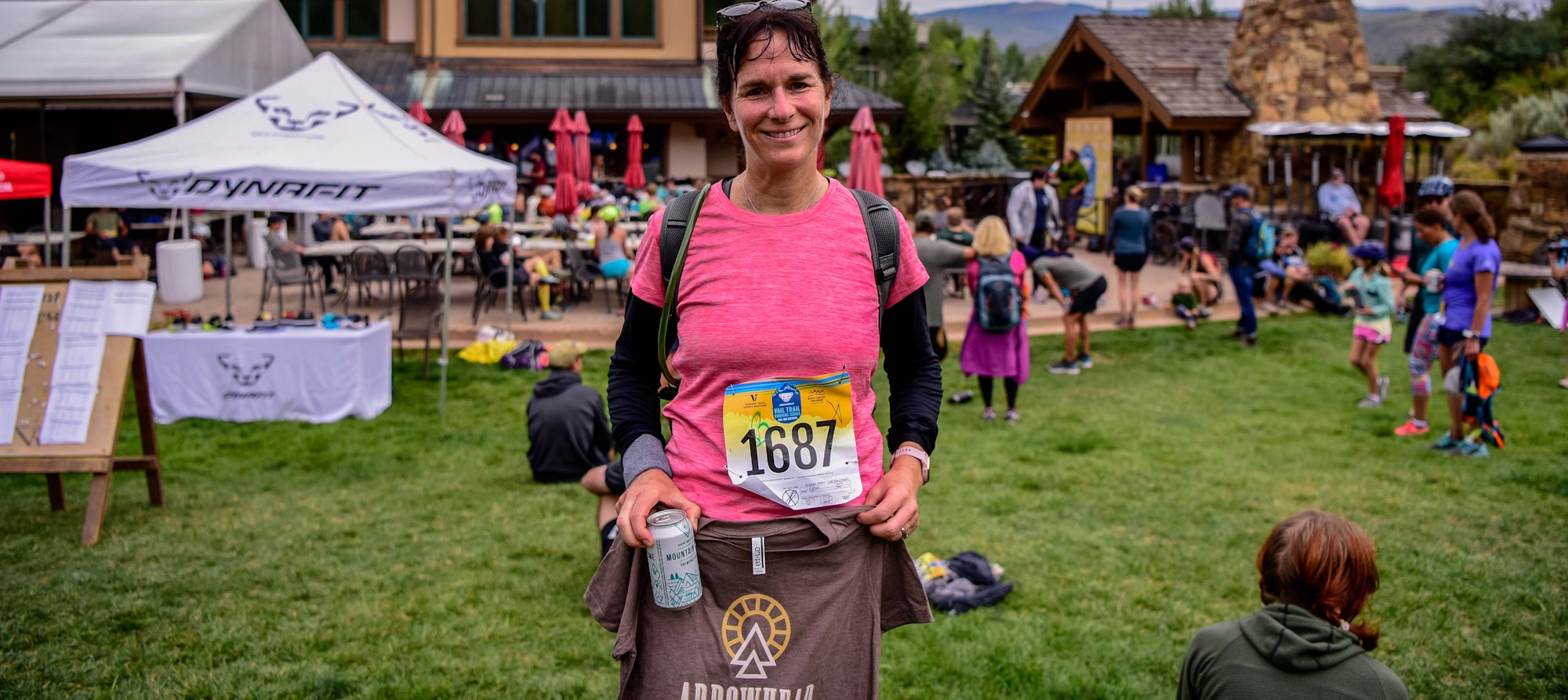 A runner with a bib number smiling at an outdoor event, holding a can. Tents and other participants in the background.