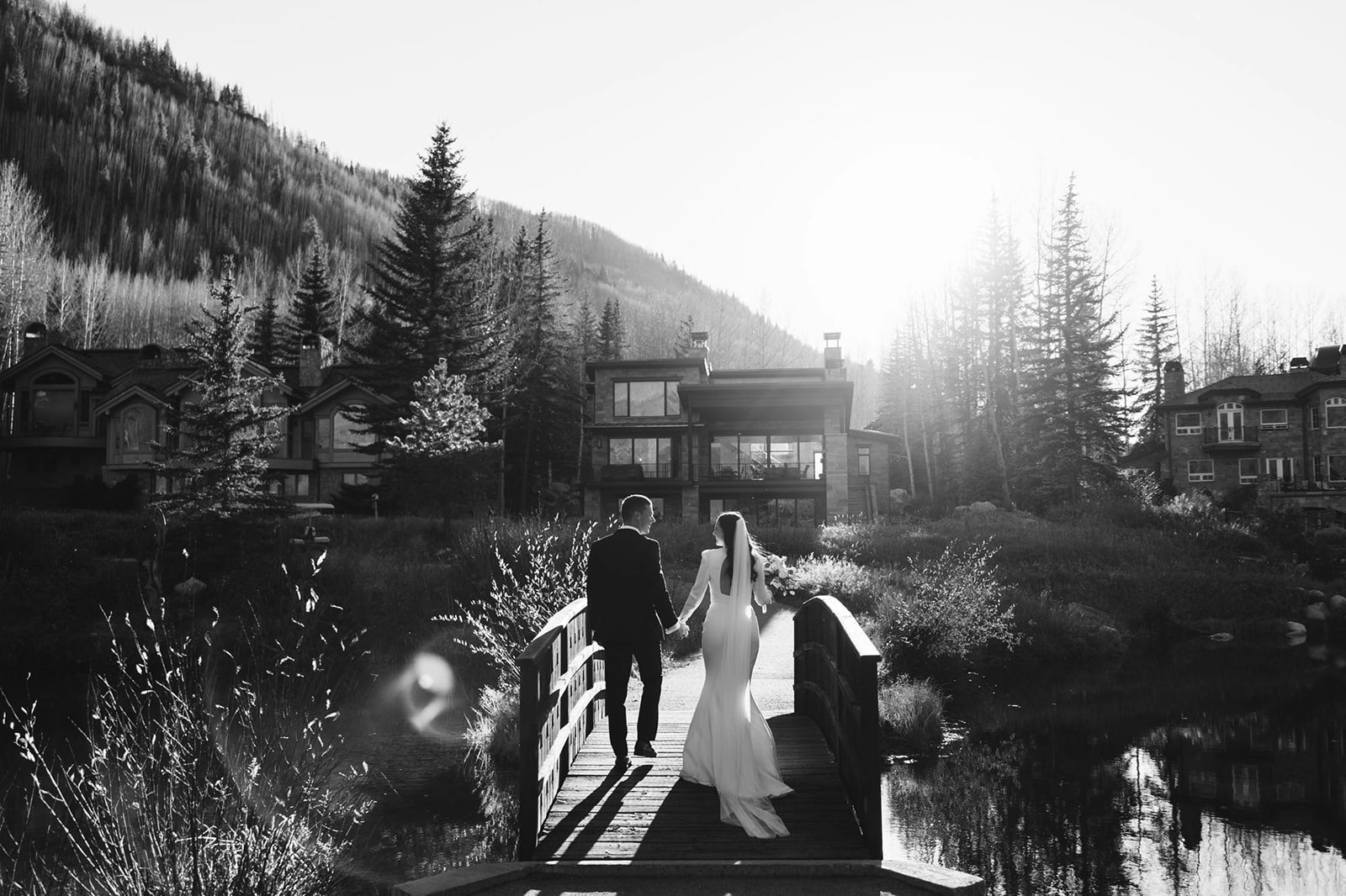 A couple in wedding attire holding hands on a bridge, with a scenic backdrop of mountains and luxury houses.