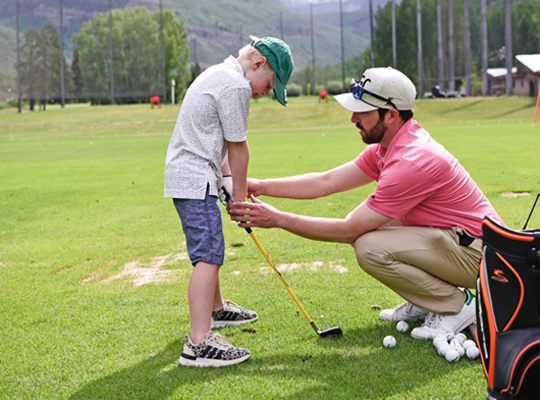 A man is teaching a young boy how to play golf on a practice range, with golf clubs and balls beside them.