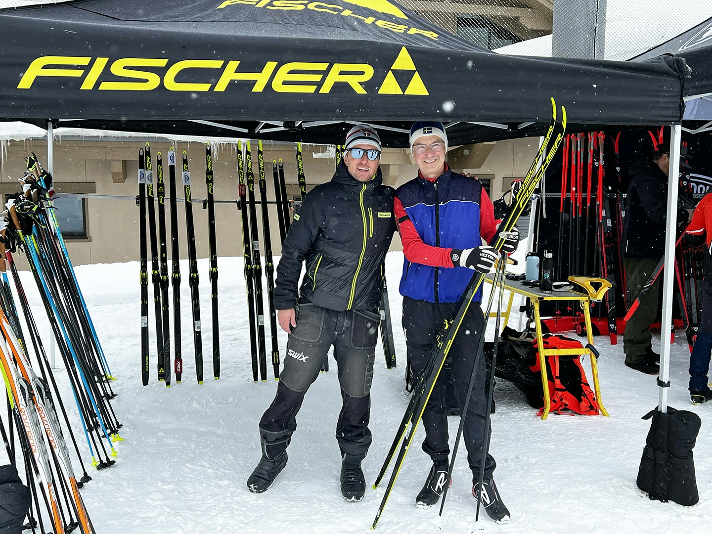 Two men standing in the snow under a tent with cross-country skis and equipment.