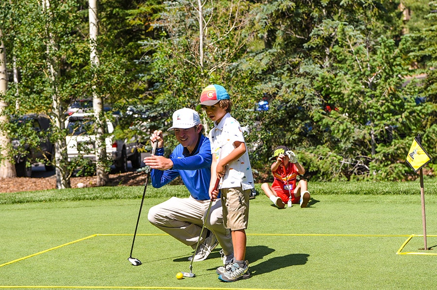 An adult instructs a child in golf putting, with a spectator in the background.