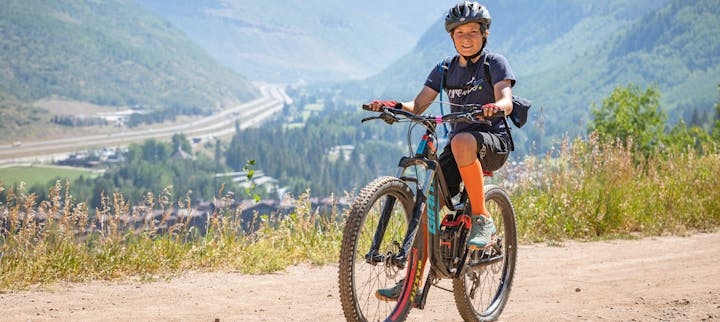 A person mountain biking on a dirt trail with a scenic backdrop of hills and clear skies.
