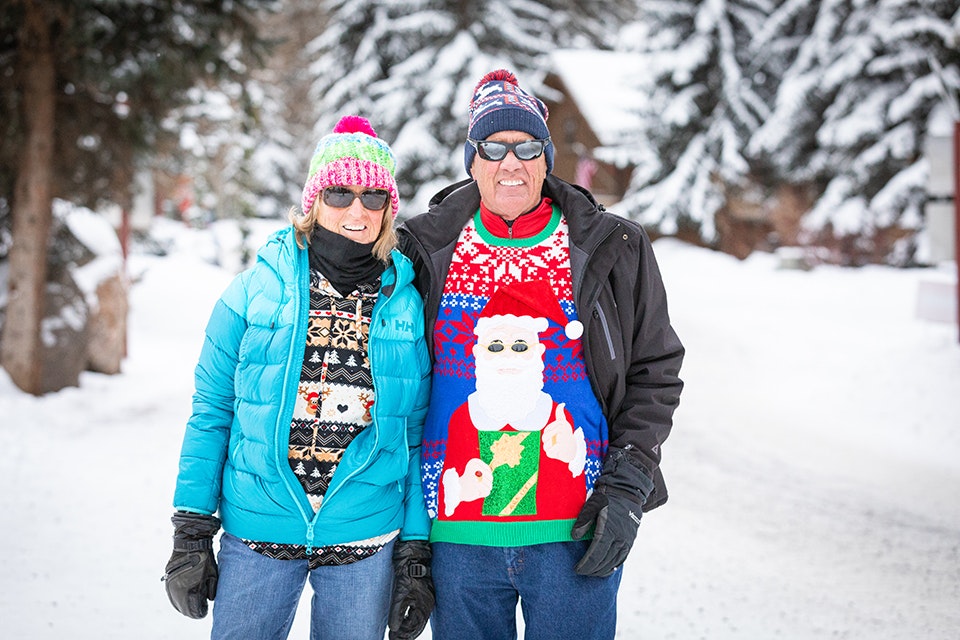 Two people in winter clothing smiling, one wearing a festive sweater, in a snowy setting.