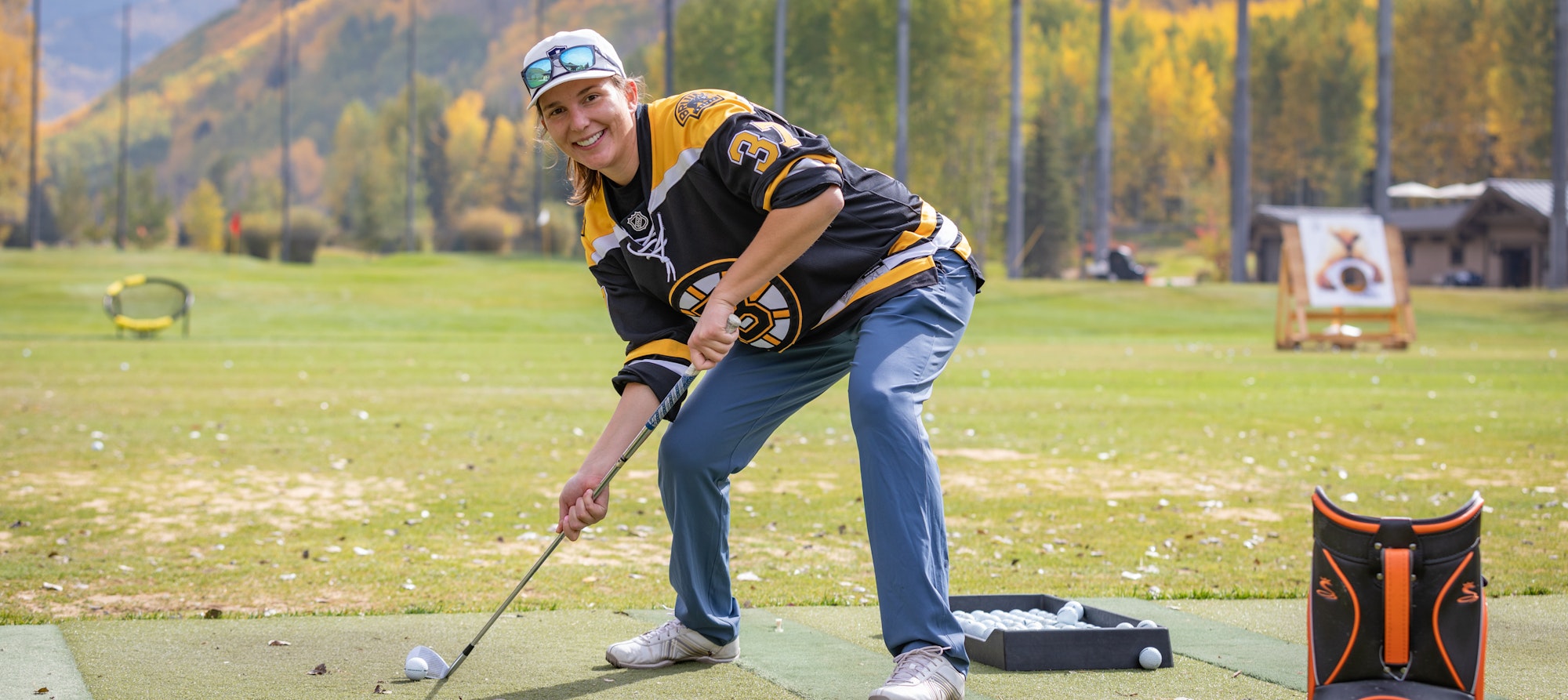 Person in a hockey jersey playing golf on a sunny day with autumn foliage in the background.