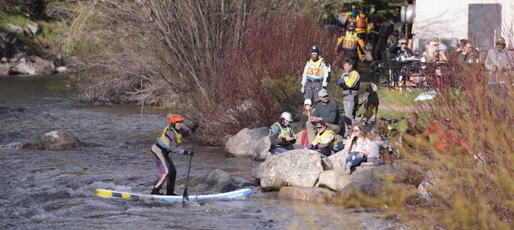 Person paddleboarding on river, spectators on shore, sunny outdoor setting.