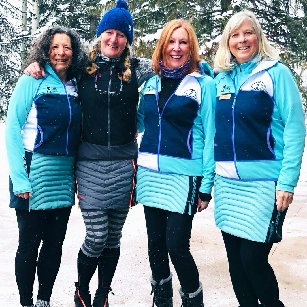 Four women in matching winter outfits smiling, with snow in the background.