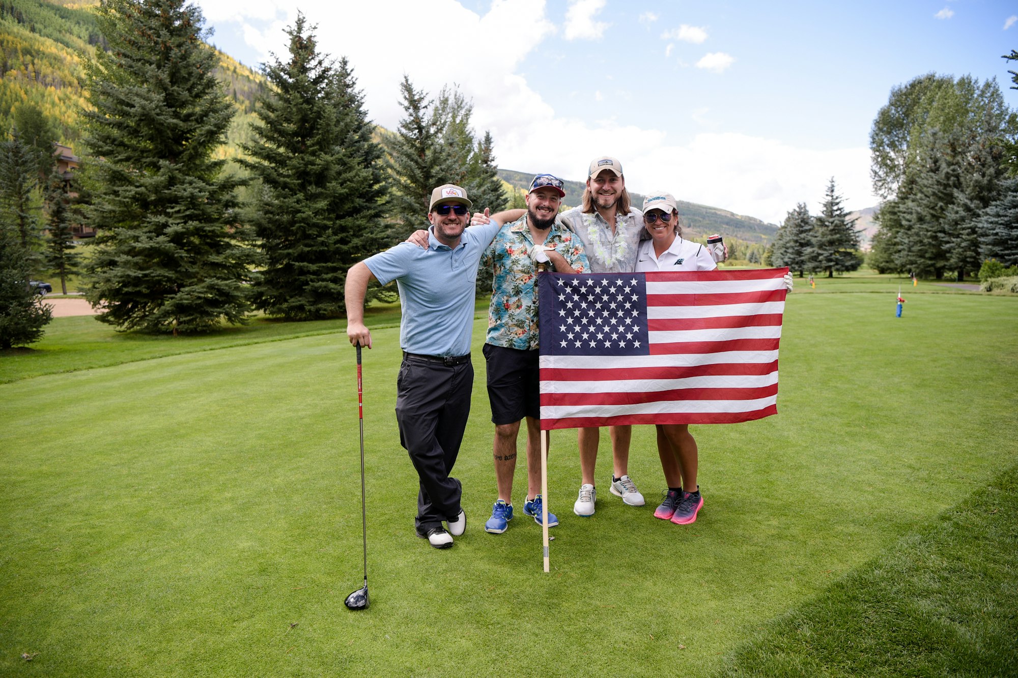 Four people holding a US flag on a golf course, smiling, in casual sportswear, surrounded by greenery.