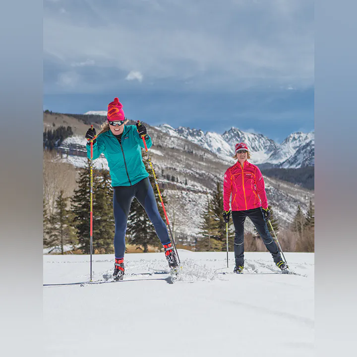 Two people cross-country skiing on snow with mountains in the background.