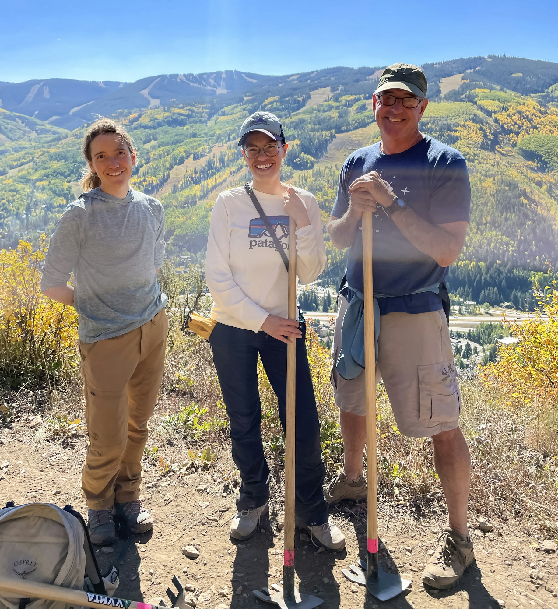 Three hikers with tools on a sunny mountain trail with autumn foliage.