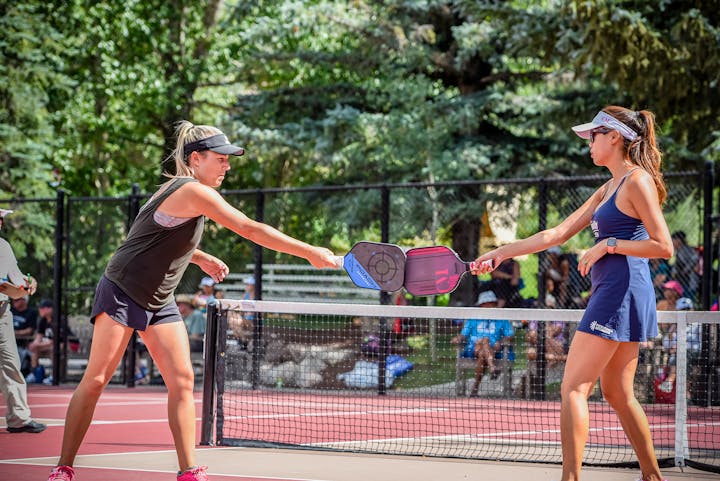 Two women touching paddles over a net, likely playing pickleball, with onlookers in the background.