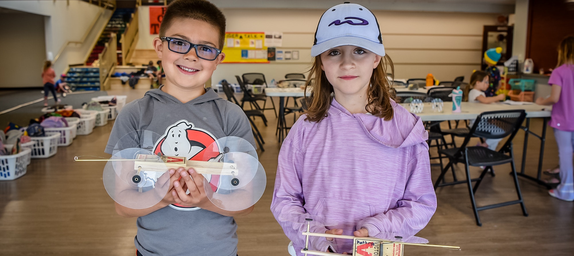 Two kids with homemade CD hovercrafts smiling in a classroom.