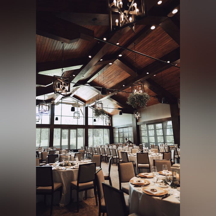 Elegant dining hall with set tables, wooden beams, chandeliers, and large windows.