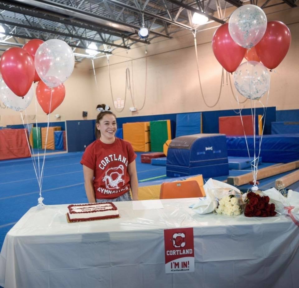 A happy young woman in a gym with balloons, a cake on a table, and "CORTLAND I'M IN!" signs.