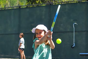 A child playing tennis, wearing a cap, and holding a racket about to hit a tennis ball, with another player in the background.