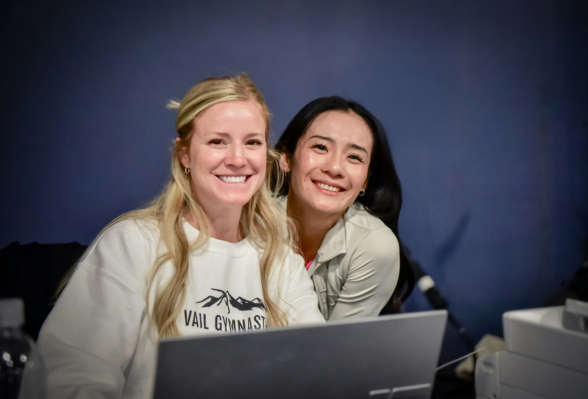 Two people smiling and posing together, with one wearing a "Vail Gymnastics" shirt, seated behind a laptop.