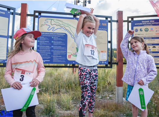 Three children with race bibs and certificates standing before informational signs, one raising her arms in a victorious pose.