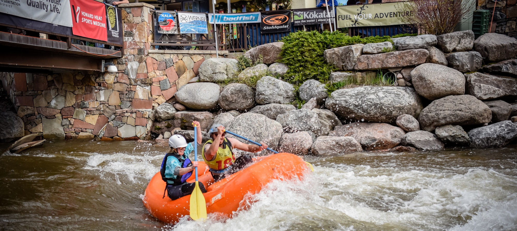 Two people whitewater rafting, navigating through rocky rapids with paddles in hand, under a bridge with outdoor banners.