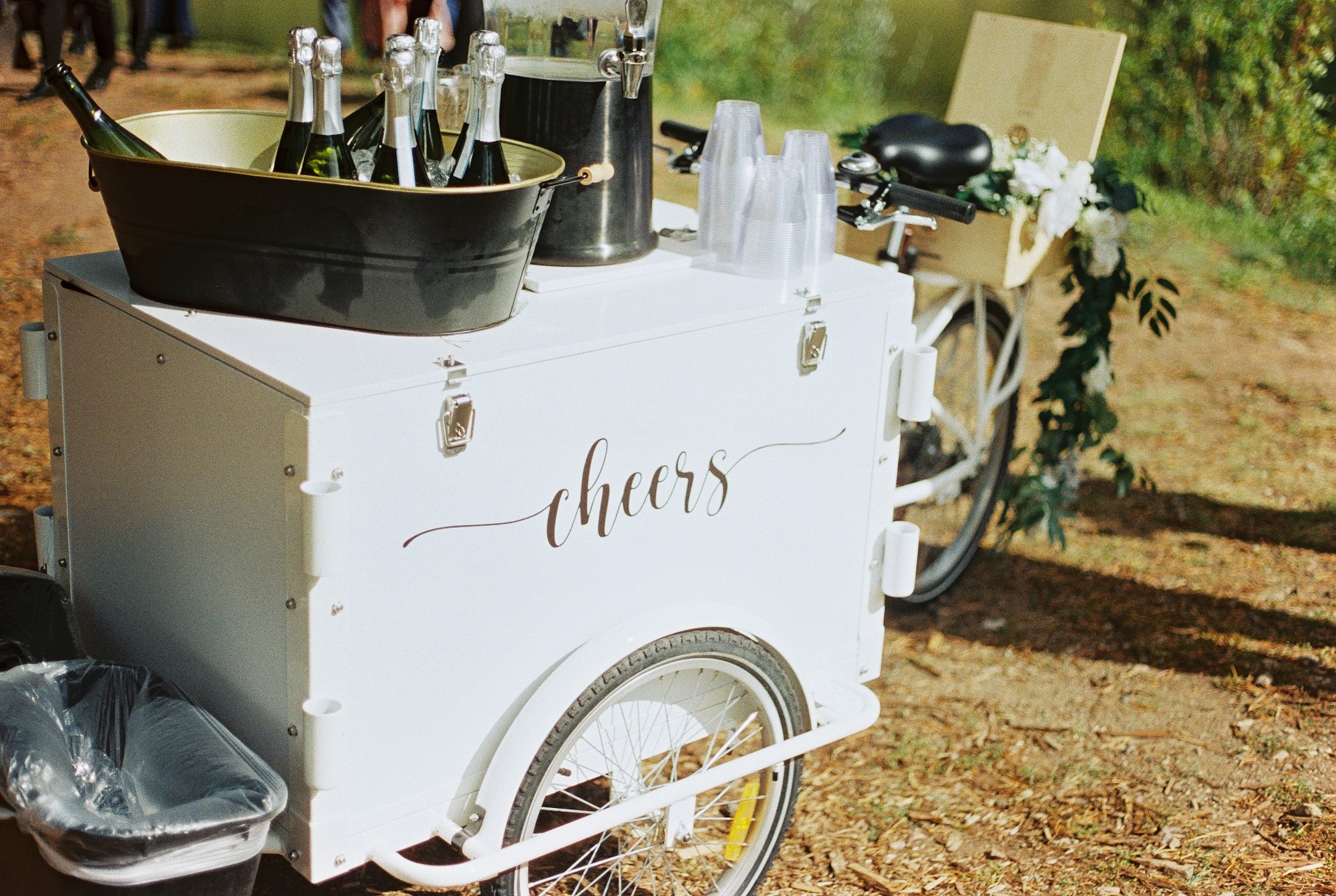 A bicycle cart with champagne bottles, glasses, and a "cheers" sign, likely at an event.