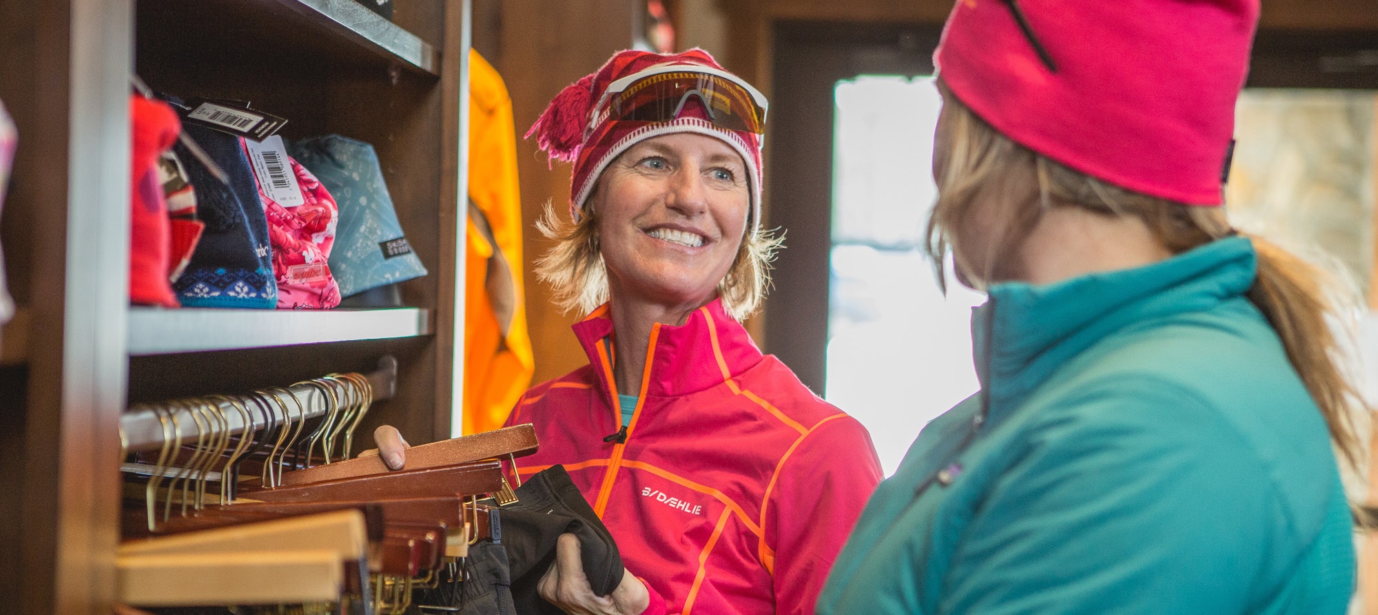 Two women in warm clothing are inside a store, one holding gloves, both smiling and interacting with each other.
