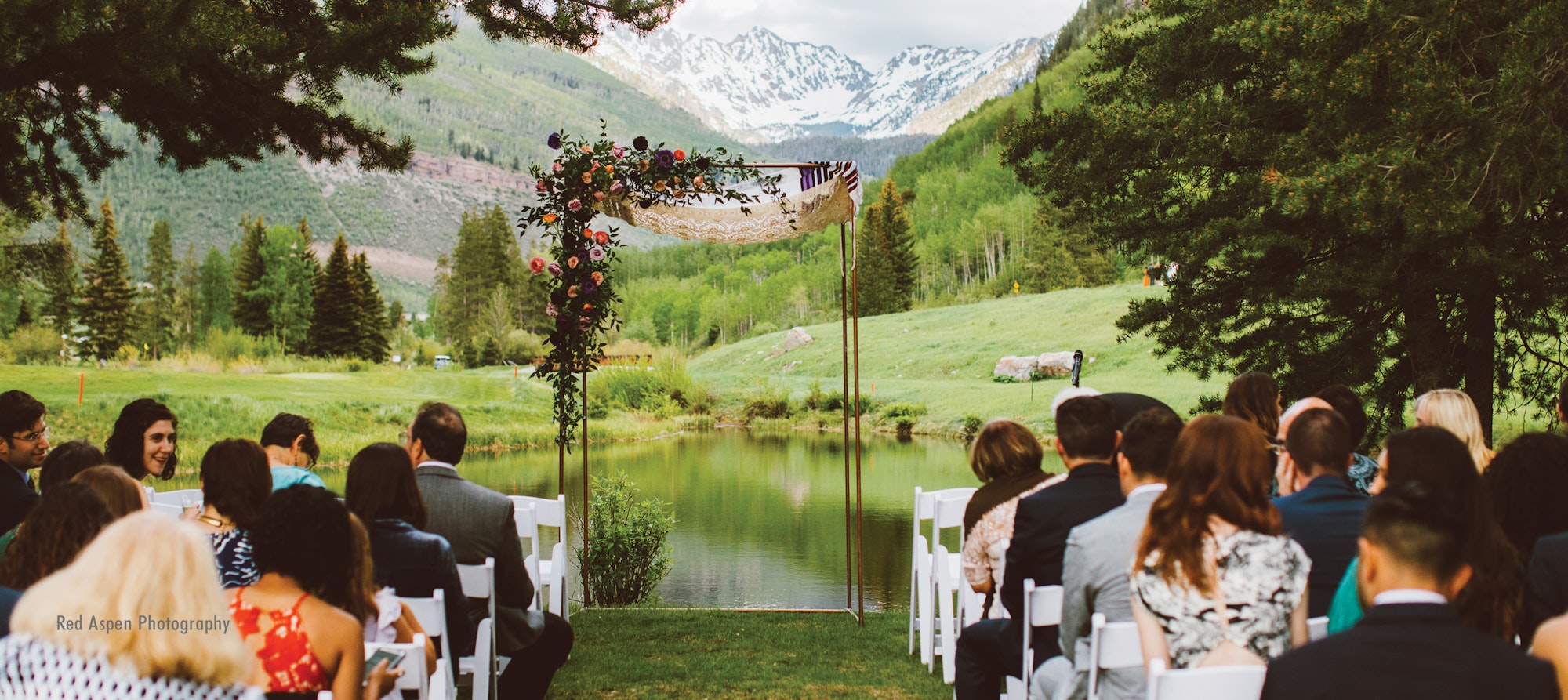 Outdoor wedding ceremony with guests, floral arch, mountain backdrop, and a pond.