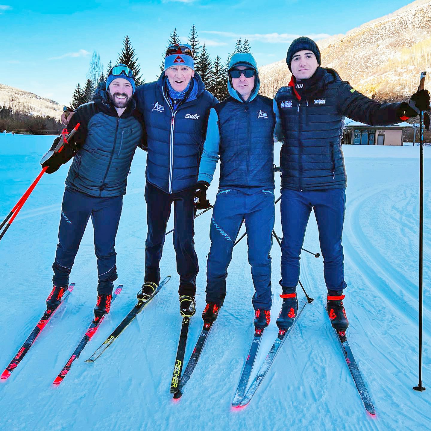 Four people wearing ski gear posing together on a snowy landscape.