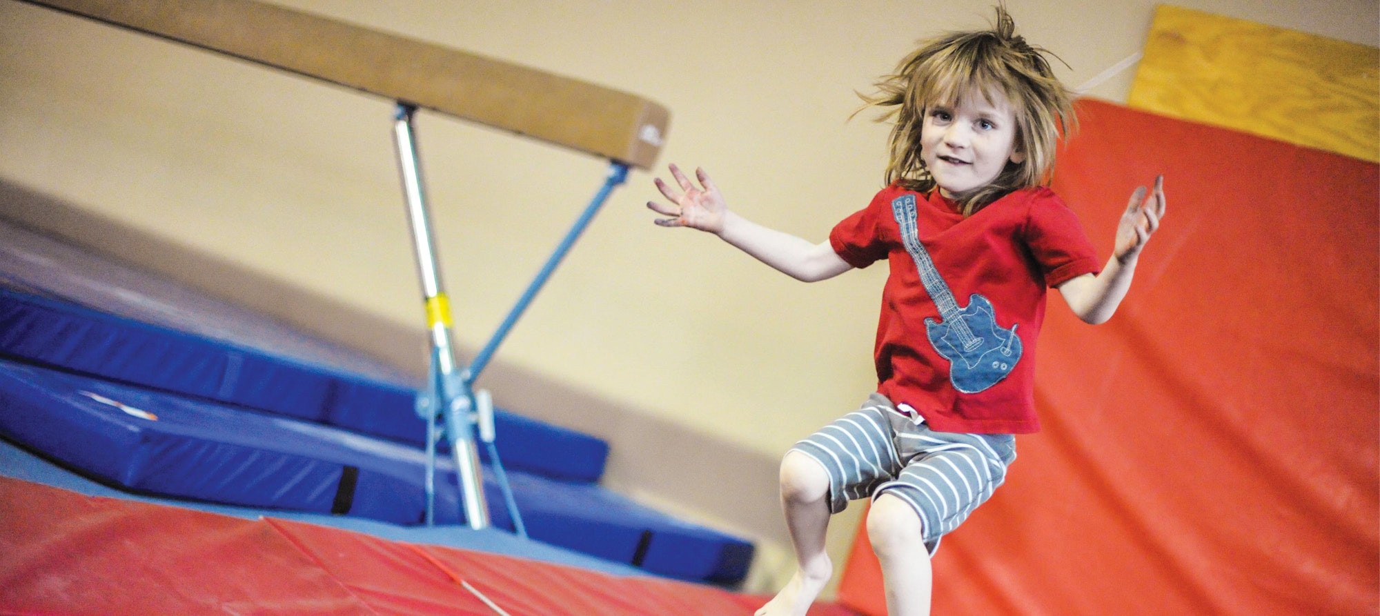 A child with tousled hair is joyfully jumping near a balance beam in a gymnasium with colorful mats.