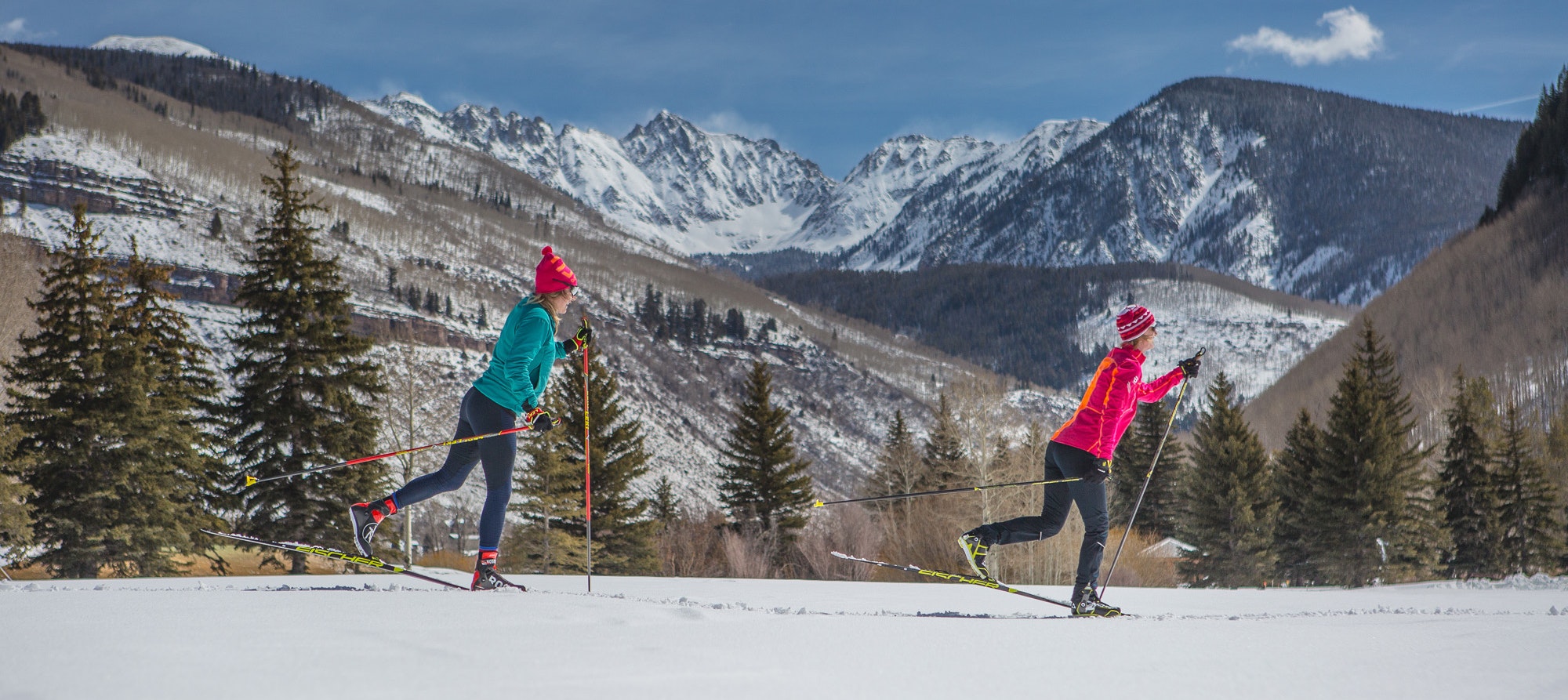 Two people cross-country skiing on a snowy landscape with mountains and trees in the background.