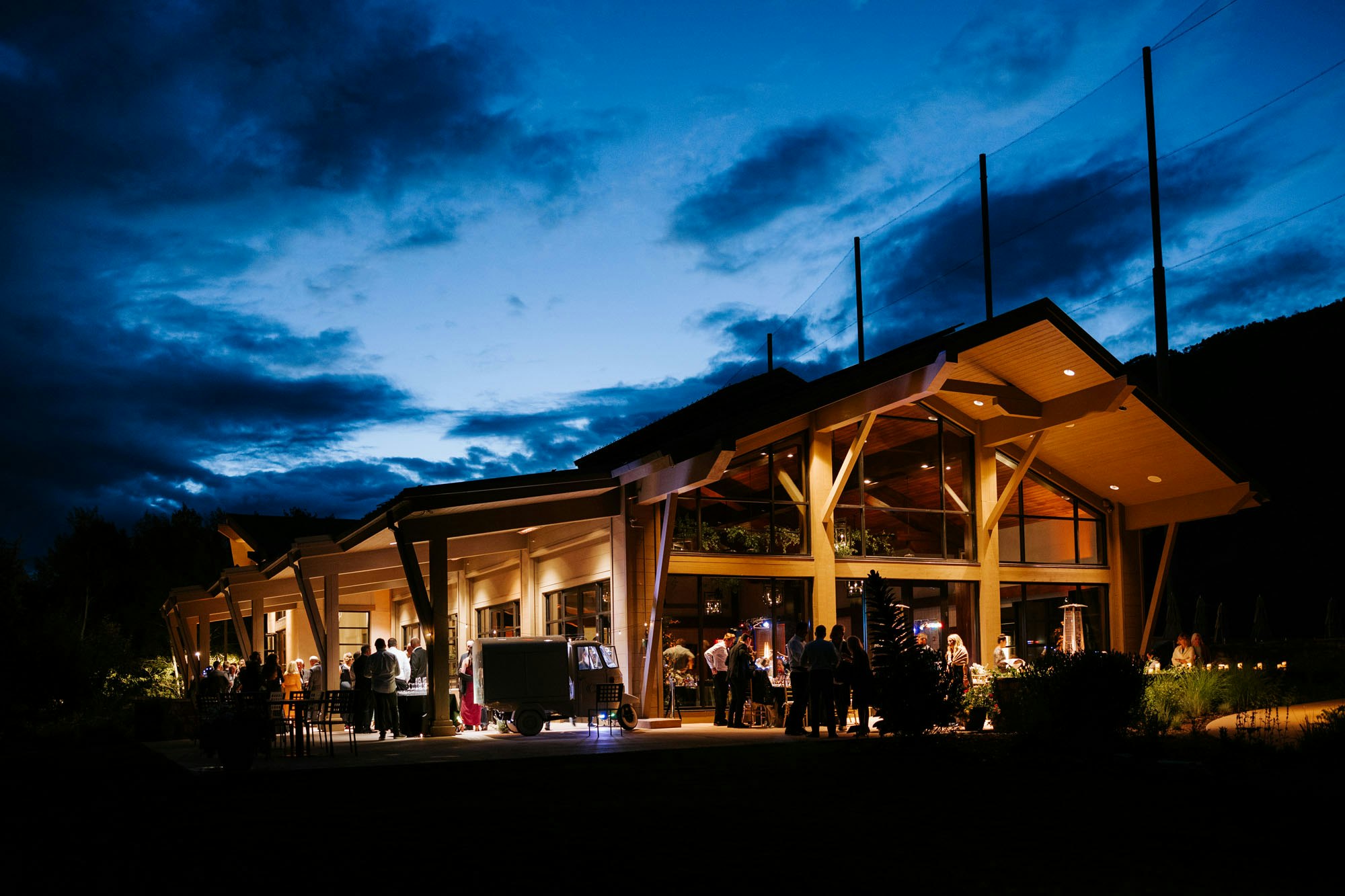 The Vail Golf & Nordic Clubhouse lit up at night, with people gathered outside, under a dramatic evening sky.