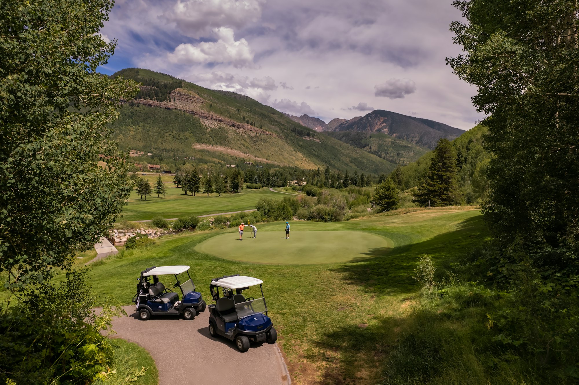 A scenic golf course with golfers and carts amidst mountains and greenery.