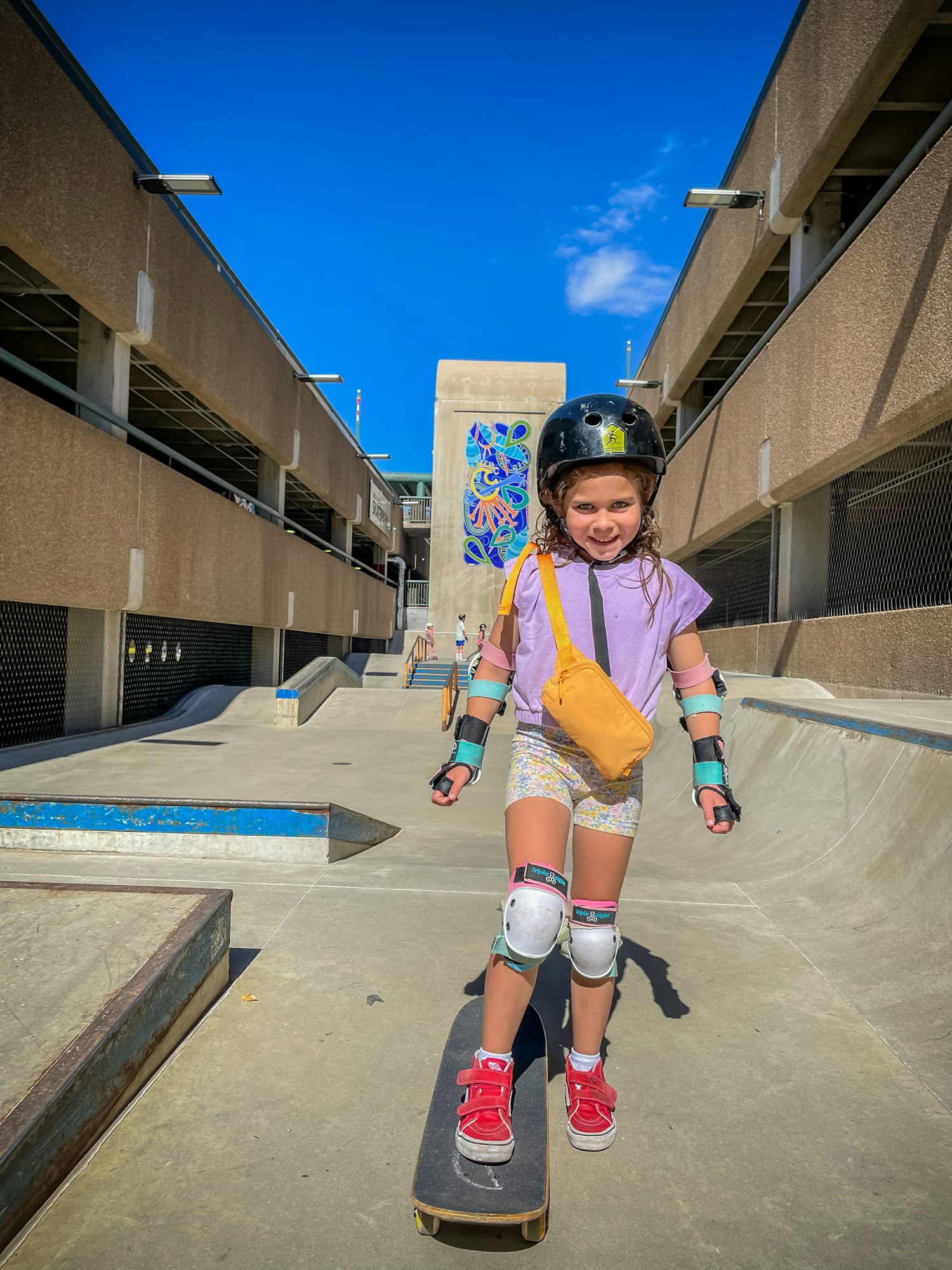 A smiling child in safety gear stands on a skateboard at a skatepark.