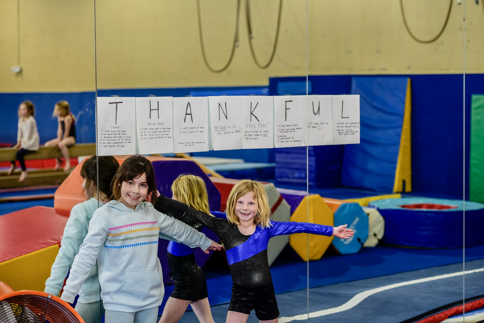 Two girls in a gym with a "THANKFUL" sign on the mirror, surrounded by colorful gym equipment.