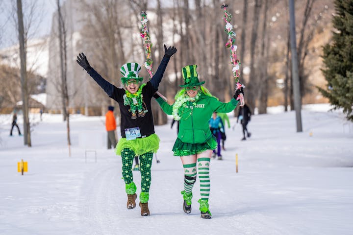 Two people in green St. Patrick's Day outfits running joyfully in the snow.