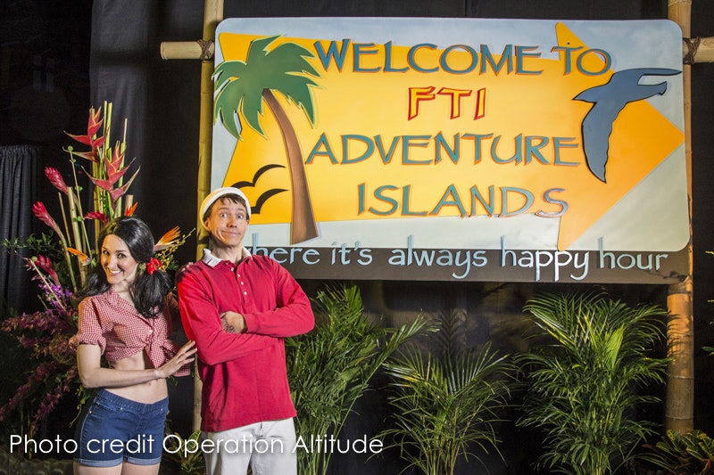 A man and a woman posing in front of a 'Welcome to Adventure Islands' sign with tropical decor.